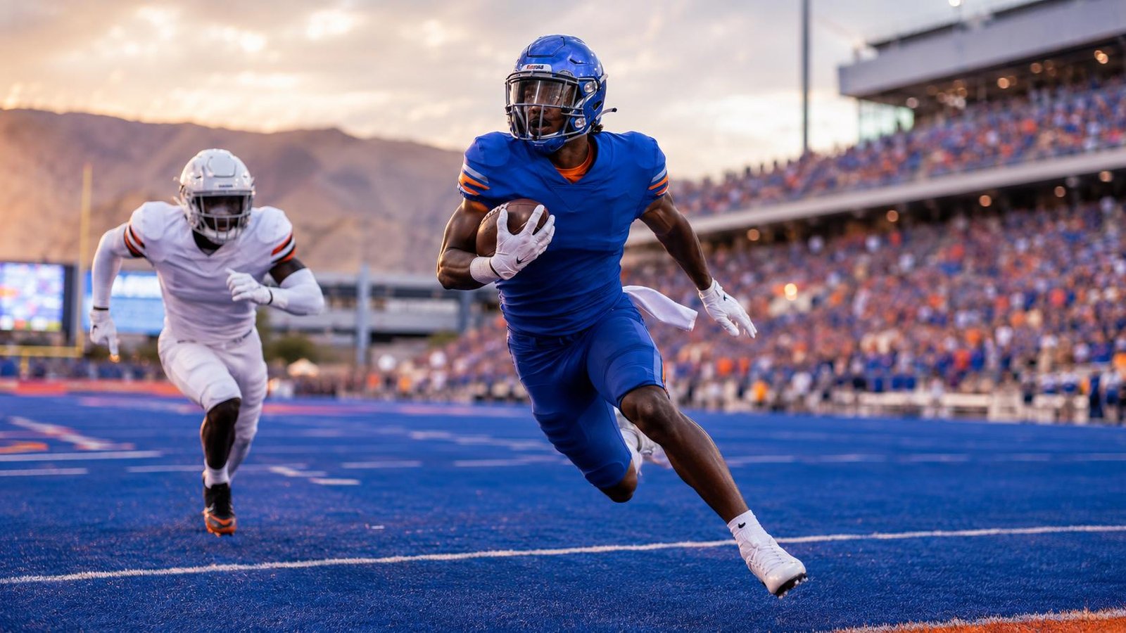 A football player in blue carrying the ball on a blue field while a defender gives chase at sunset.